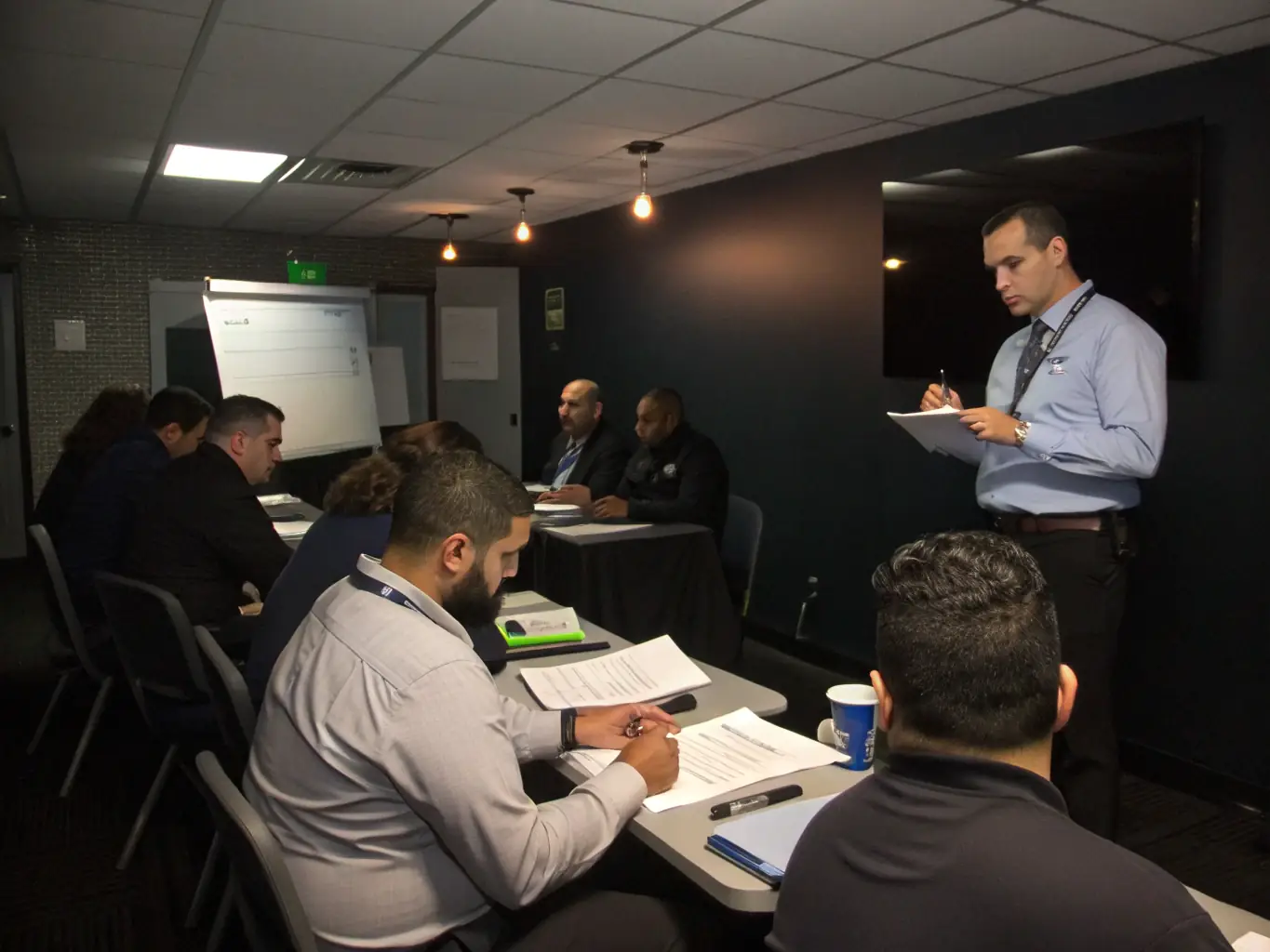 A modern training room with business professionals participating in a security awareness session, an instructor presenting insider-threat scenarios, and compliance documentation visible on the table.